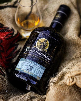 A bottle of Bunnahabhain Stuidreadair single malt Scotch whisky, covered in condensation, resting on a rustic burlap fabric next to a glass of the golden whisky and a red patterned cloth.