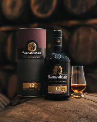 A bottle of Bunnahabhain 12 Year Single Malt Scotch whisky stands on a wooden surface next to a glass of the amber liquid, with its cylindrical packaging box beside it. The background shows blurred wooden whisky barrels.