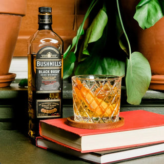 A bottle of Bushmills Black Bush Irish Whiskey sits by a whiskey glass garnished with orange peel on a coaster atop a closed book, surrounded by green potted plants, evoking notes of sherry sweetness and dried fruit.