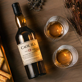 A bottle of Caol Ila 12 Year Old Single Malt Scotch Whisky rests on a wooden table with two whisky-filled glasses, dried herbs, and breadsticks, the signature peat smoke blending subtly with malt sweetness.