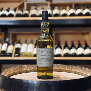 A bottle of Caol Ila Moch Single Malt Scotch Whisky rests on a wooden barrel, hinting at peat smoke and vanilla aromas, while shelves of assorted wines blur in the background, evoking a refined liquor store atmosphere.