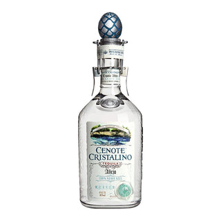 A clear glass bottle of Cenote Cristalino Añejo Tequila 700ml features a blue decorative cap and a label with a cenote illustration, product details, and notes of agave, vanilla, and toasted oak.