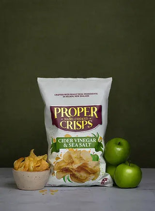A bag of Proper Crisps Cider Vinegar and Sea Salt Chips stands upright with a bowl of chips and two green apples, all set against a simple dark green background.