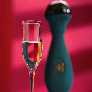 A tall, elegant glass with clear Clase Azul Mezcal Guerrero sits before a blurred teal bottle with a gold emblem, all against a vibrant pink background—capturing the mezcal’s subtle honeyed sweetness in every sip.