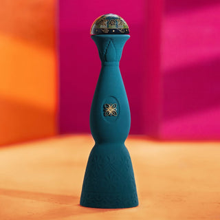 A tall, elegant blue bottle of Clase Azul Mezcal Guerrero with floral patterns and a round stopper sits on an orange surface, echoing smoky agave notes against vivid pink and orange background walls.
