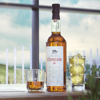 A bottle of Clynelish 14 Year Old Single Malt Whisky rests on an outdoor table beside whisky glasses, its sweet malt and citrus notes echoing the sea salt breeze of the scenic backdrop.