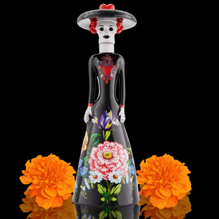 A Day of the Dead (Dia de los Muertos) sugar skull style bottle or vase with a black body, decorated with colorful floral patterns and a white hat with red flowers, flanked by two orange marigolds on a black background. Doña Celia Tequila Blanco 750ml