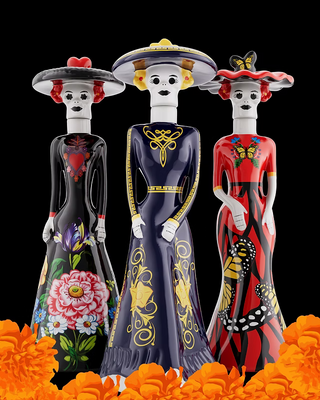 Three decorative Day of the Dead (Día de los Muertos) figurines shaped like women in long dresses, standing on a black background with orange marigolds at the bottom. The left figurine wears a black dress with a pink flower and red hat; the center figurine wears a navy blue dress with gold patterns and a yellow hat; the right figurine wears a red dress with black and gold patterns and a red hat. Doña Celia Tequilas.