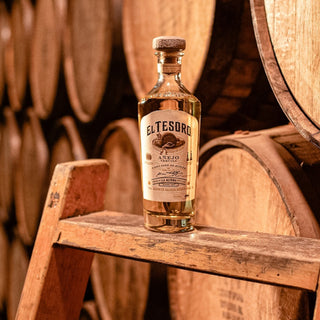 A bottle of El Tesoro Añejo Tequila 750ml rests on a wooden ladder, with barrels stacked behind it, evoking the distillery where aromas of cooked agave and toasted oak fill the air.