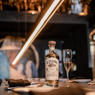 A bottle of El Tesoro Añejo Tequila 750ml and a filled champagne flute rest on a dark restaurant table set for dining, warmly lit by a pendant light amid a softly blurred background.