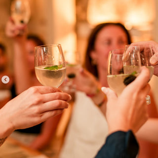 A group toasts with wine glasses filled with El Tesoro Añejo Tequila 750ml, each garnished with fresh mint. The air hints of cooked agave, while a warm, festive glow lights up the background.