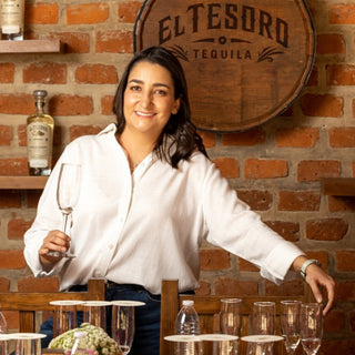 A woman in a white blouse smiles with a wine glass, standing before a brick wall and an El Tesoro Reposado Tequila 750ml barrel. Notes of toasted oak and sweet agave fill the air around her tasting table.