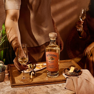 A person holds a wine glass beside a bottle of El Tesoro Reposado Tequila 750ml, its cooked agave and toasted oak notes pairing with cheese and chocolate on a wooden tray; another person sits in the background holding a drink.