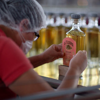 Wearing a hairnet, glasses, and a mask, a worker carefully labels a bottle of El Tesoro Reposado Tequila 750ml, with several identical bottles arranged in the background.