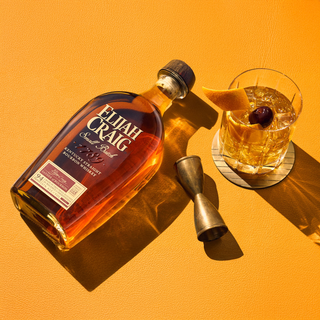 Bottle of Elijah Craig bourbon with a cocktail glass and jigger on an orange background