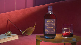 A bottle of Four Pillars Single Highland Single Malt Scotch whisky stands on a small gold table next to a glass of the dark amber liquid, set against a red velvet sofa with a book and decorative leaf in the background.