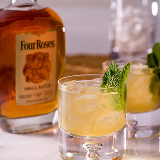 A glass of bourbon cocktail with ice and mint sits before a Four Roses Small Batch Kentucky Straight Bourbon Whiskey bottle, known for caramel and baking spice notes, with another drink and a glass container blurred in the background.