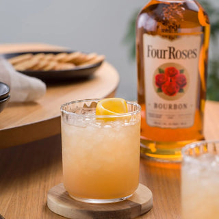 A glass of iced bourbon cocktail, layered with spice and vanilla notes, sits on a wooden coaster garnished with a lemon twist. Behind it are crackers and a bottle of Four Roses Kentucky Straight Bourbon Whiskey, creating an inviting scene.