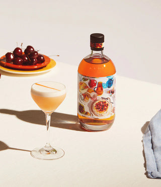 Bottle of alcoholic beverage with a colorful label next to a glass and cherries on a white surface