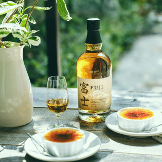 A bottle of Fuji Single Malt Japanese Whisky, a glass of whisky, and two crème brûlées rest on a wooden table beside a white vase with green leaves. Sunlight bathes the scene, while hints of white peach evoke warmth and invitation.