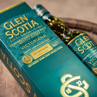 A bottle of Glen Scotia Victoriana Single Malt Scotch Whisky sits beside its matching green and gold box on a wooden surface. The label suggests notes of dark chocolate and caramelised fruits, with the branding clearly shown.