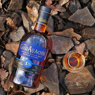 A bottle of GlenAllachie 15 Year Old Single Malt Scotch Whisky rests on rocks and dried leaves beside a whisky glass, inviting you to enjoy its floral honey character and sweet spice layers.