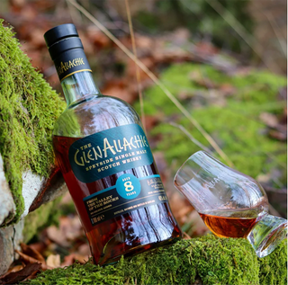 A bottle of GlenAllachie 8 Year Old Single Malt Scotch Whisky rests on mossy rocks outdoors beside a glass of its amber spirit, suggesting hints of cinnamon and rich dark chocolate.