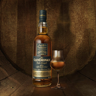 A bottle of Glendronach Cask Strength Batch 10 Single Malt Scotch Whisky stands next to a filled whisky glass on a wooden surface, set against rustic wood paneling and evoking inviting hints of dried fruits and sherry sweetness.