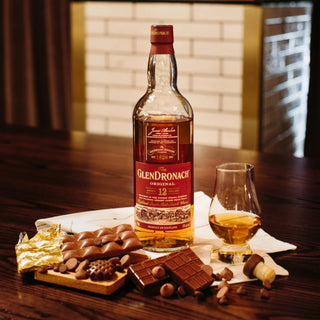 A bottle of Glendronach 12 Year Old Single Malt Scotch Whisky rests on a wooden table with a whisky glass, unwrapped chocolate bar, dark chocolate pieces, and cork, evoking creamy vanilla notes against a white-tiled backdrop.