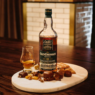 A bottle of Glendronach 15 Year Old Revival Single Malt Scotch Whisky sits by a filled glass, surrounded by dark chocolate, dried fruits, and nuts on marble atop wood—showcasing its signature rich sherry cask influence.