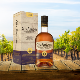 A bottle of GlenAllachie 11 Year Old Grattamacco Cask Finish Single Malt Scotch Whisky and its box sit on a wooden surface, set against a scenic vineyard backdrop—inside are notes of caramel and dark chocolate.