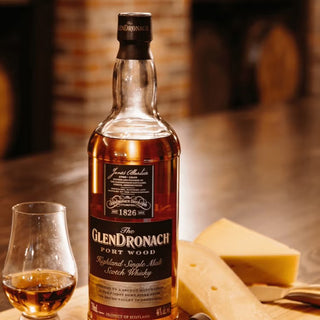 A bottle of Glendronach Port Wood Single Malt Scotch Whisky rests on a wooden table beside a glass and cheese, as aromas of ripe berries and port wine drift through the softly blurred brick fireplace in the background.