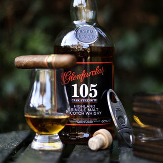 A bottle of Glenfarclas 105 Cask Strength Single Malt Scotch Whisky sits behind a whisky glass filled with amber liquid, alongside a cigar, cigar cutter, and cork on an oak surface.