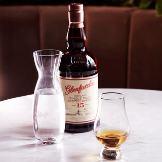 A bottle of Glenfarclas 15 Year Old Single Malt Scotch Whisky sits on a marble table next to a carafe and a whisky glass, offering rich, full-bodied flavor and deep malty tones with every sip.