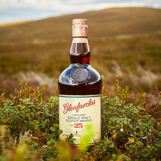 A bottle of Glenfarclas 25 Year Old Single Malt Scotch Whisky stands upright in grassy countryside with rolling hills behind it, evoking notes of dried fruits and dark chocolate beneath a partly cloudy sky.