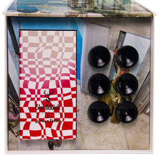A Glenfiddich Grand Cru 23 Year Old x Jordan Dalah Box Set with a wavy red-and-white checkered design sits beside six black round objects, evoking sandalwood and peach notes; a woman in gold appears behind a glass wall.