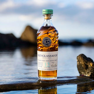 A bottle of Glenglassaugh 12 Year Old Single Malt Scotch Whisky rests on a wet rock near water, with a blurred coastal background and rocks, evoking notes of ripe apples and pears.