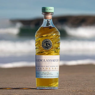 A bottle of Glenglassaugh Sandend Single Malt Scotch Whisky rests on a sandy shore, evoking notes of salted caramel and tropical fruit as blurred sea waves and rocky outcrops frame the background.