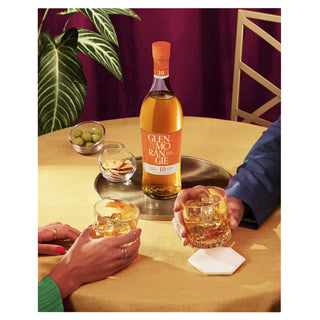 A bottle of Glenmorangie 10 Year Old Single Malt Scotch Whisky rests on a round tray with two whisky glasses, olives, and crackers atop a yellow tablecloth as two people reach for their drinks.