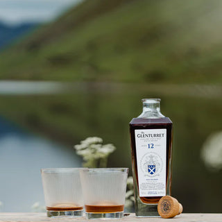 A bottle of Glenturret 12 Year Old Single Malt Scotch Whisky stands beside two partially filled glasses and a cork on a wooden surface outdoors, with greenery and water blurred in the background, evoking vanilla and gentle oak spice.