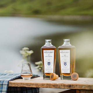 A bottle of Glenturret 7 Year Old Peat Smoked Single Malt Scotch Whisky sits on a wooden table by a lake, next to a glass of smoky whisky, corks, and a blue tartan cloth draped over the table.