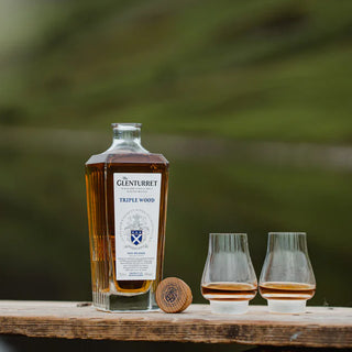 A bottle of Glenturret Triple Wood Single Malt Scotch Whisky stands on a wooden surface beside two partially filled glasses, with a cork in front. Blurred greenery hints at the whisky’s toasted oak and dried fruit notes in the background.