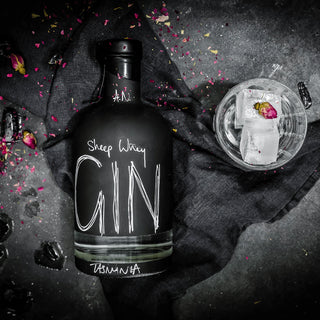 A black bottle of Gin with white handwritten-style text reading 'Sheep Waxy GIN' and 'HUMMA' near the base, arranged on a dark cloth beside a clear glass with ice and a rose petal, on a dark textured surface with scattered flower petals.