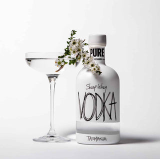 A clear glass coupe coupe glass with a clear liquid and a small white blossom sprig, next to a frosted bottle labeled 'PURE VODKA' and 'Shop Whey VODKA TASMANIA' on a clean white background.