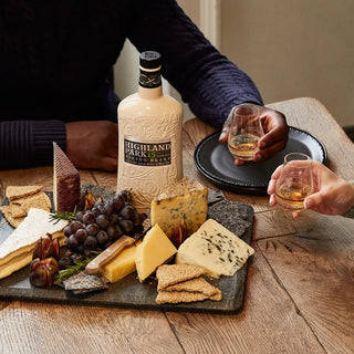 A slate board with cheeses, crackers, grapes, and honey sits on a wooden table. Two people toast glasses of Highland Park 15 Year Old Viking Heart Single Malt Scotch Whisky, its peat smoke and sherry notes enhancing the cozy moment.