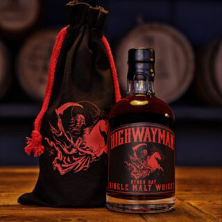 A bottle of Highwayman Crawling King Snake Single Malt Australian Whisky with a black and red skeleton label sits beside a matching drawstring bag on wood, barrels blurred in the background, with earthy peat hints.
