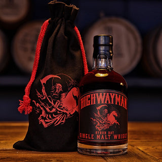 A bottle of Highwayman Far Beyond Peatera Single Malt Australian Whisky with a black and red label and skeleton illustration sits beside a matching drawstring bag on wood, with peaty aromas and barrels in the background.