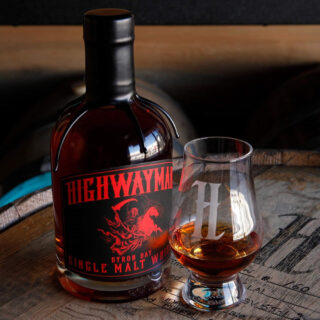 A bottle of Highwayman The Indiana Gentleman Single Malt Australian Whisky with a red and black label sits beside a whisky glass bearing a white H logo, both on a wooden barrel. The amber liquid suggests caramel fudge and earthy campfire smokiness.