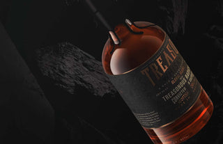 A close-up of a bottle of Highwayman Treacherous Radicals Single Malt Australian Whisky against a dark, rocky background, with warm lighting showcasing the amber liquid and embossed label, evoking mocha and roasted mixed nut notes.