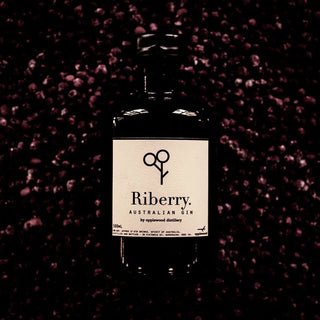 A dark glass bottle of Applewood Riberry Gin is centered among scattered dark berries. The minimalist label, featuring a small scissors-like graphic, highlights native botanicals and aromatic spice from Applewood Distillery.